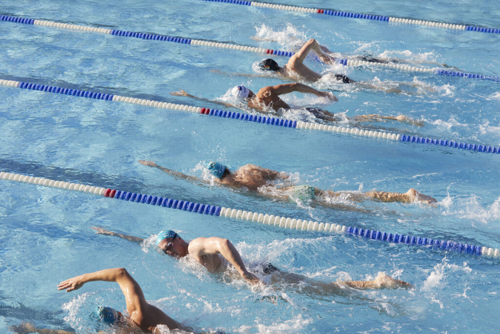 Photo looking downwards upon five adults across several lap lanes, swimming beside each other in freestyle stroke.