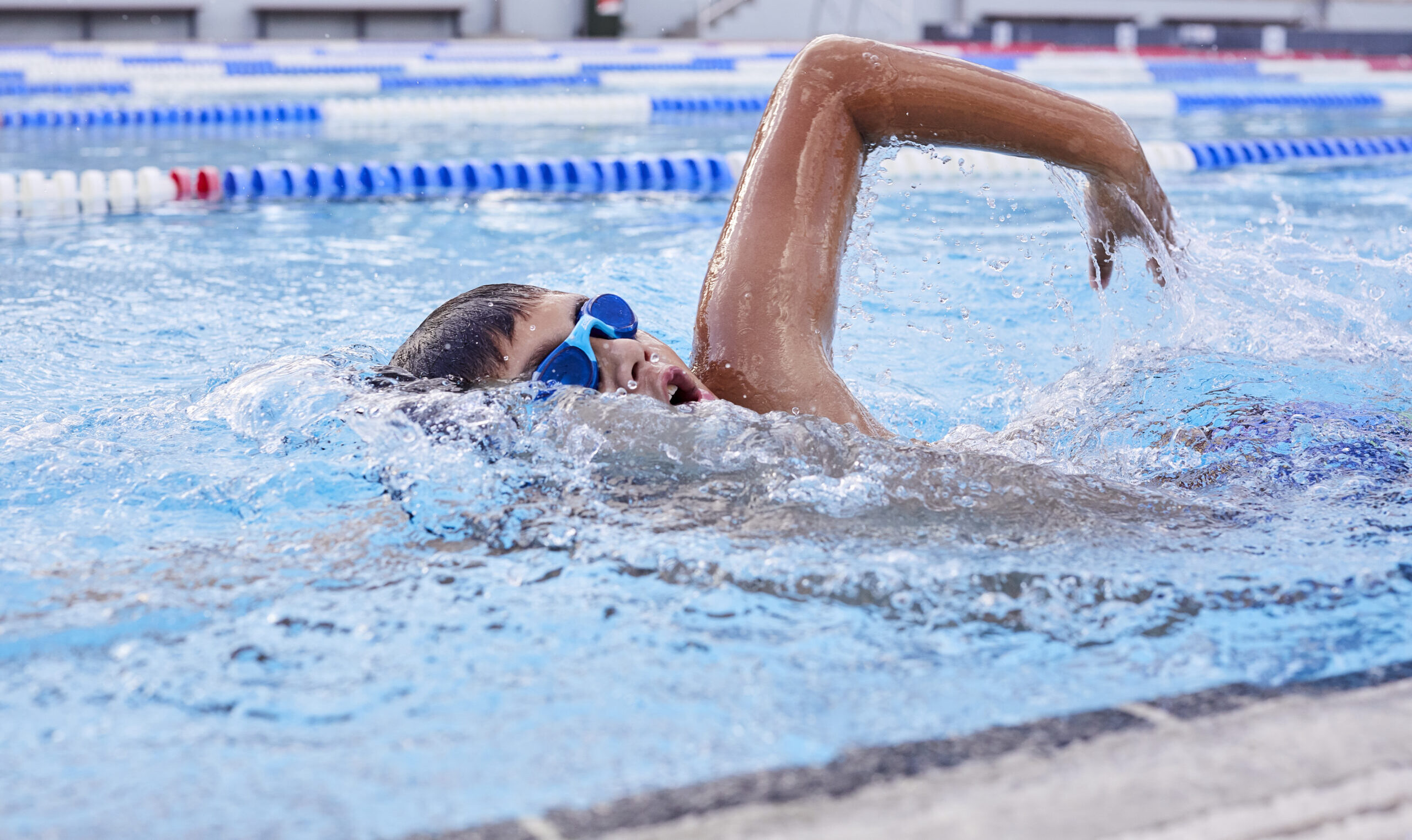 Close up photo of a young boy swimming freesytle in a lap swimmng pool, wearing blue goggles and taking a breath above the water.